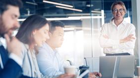 Selbstbewusste Frau im Business-Outfit steht vor einem Team am Besprechungstisch – Sinnbild für Führungskompetenz, strategisches Denken und professionelles Immobilienmanagement.