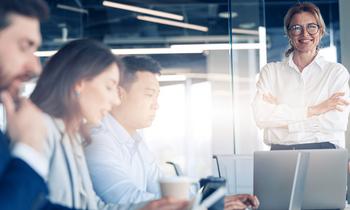 Selbstbewusste Frau im Business-Outfit steht vor einem Team am Besprechungstisch – Sinnbild für Führungskompetenz, strategisches Denken und professionelles Immobilienmanagement.
