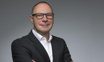Erwin Hesser, MBA-Absolvent der FH Burgenland, blickt selbstbewusst in die Kamera. Er trägt einen dunklen Anzug und eine Brille, der Hintergrund ist neutral grau gehalten.
