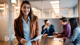 Junge Frau mit Unterlagen in der Hand steht selbstbewusst in einem modernen Besprechungsraum. Im Hintergrund diskutiert ein kleines Team – sinnbildlich für psychologisches Know-how und innovative Ansätze in der neuen Arbeitswelt, wie sie im DAS Wirtschaftspsychologie und New Work vermittelt werden.