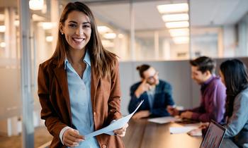 Junge Frau mit Unterlagen in der Hand steht selbstbewusst in einem modernen Besprechungsraum. Im Hintergrund diskutiert ein kleines Team – sinnbildlich für psychologisches Know-how und innovative Ansätze in der neuen Arbeitswelt, wie sie im DAS Wirtschaftspsychologie und New Work vermittelt werden.