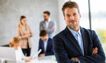 Selbstbewusster Mann im Business-Outfit steht mit verschränkten Armen vor einem modernen Bürofenster. Im Hintergrund arbeiten mehrere Personen an Laptops und Tablets. Das Bild symbolisiert den MBA Digital Business für Akademiker*innen an der FH Burgenland.