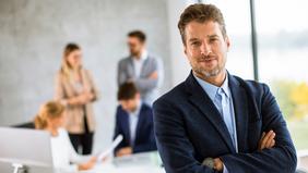 MBA Digital Business Selbstbewusster Mann im Business-Outfit steht mit verschränkten Armen vor einem modernen Bürofenster. Im Hintergrund arbeiten mehrere Personen an Laptops und Tablets. Das Bild symbolisiert den MBA Digital Business für Akademiker*innen an der FH Burgenland.