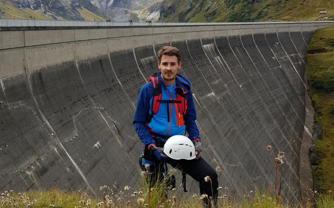 Constantin Haider in Outdoor-Ausrüstung mit Helm vor einem großen Staudamm in Berglandschaft.