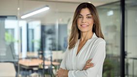 MBA Controlling Frau in weißer Bluse steht selbstbewusst mit verschränkten Armen in einem modernen Büro mit Glaswänden. Sie symbolisiert die Zielgruppe des MBA Controlling der FH Burgenland, der Absolvent*innen auf Führungspositionen vorbereitet.