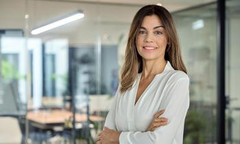 Frau in weißer Bluse steht selbstbewusst mit verschränkten Armen in einem modernen Büro mit Glaswänden. Sie symbolisiert die Zielgruppe des MBA Controlling der FH Burgenland, der Absolvent*innen auf Führungspositionen vorbereitet.