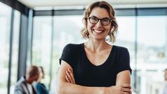 Selbstbewusste Frau mit Brille und verschränkten Armen in lichtdurchflutetem Raum – steht für Führungsstärke, Kommunikationskompetenz und berufliche Weiterbildung im Fernstudium.