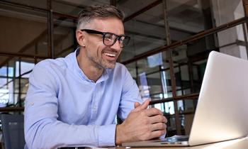MBA Digitale Geschäftsmodelle und Geschäftsmodellentwicklung Lächelnder Mann mit Brille und hellem Hemd arbeitet konzentriert an einem Laptop – Sinnbild für flexibles Online-Studium im Bereich digitale Geschäftsmodelle an der Hochschule Burgenland.