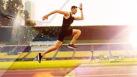 Sportlicher Mann im schwarzen Outfit sprintet über eine Laufbahn in einem Stadion bei Sonnenlicht. Im Hintergrund sind Tribünen und Kameraequipment sichtbar – symbolisch für das praxisorientierte MSc-Programm Sportphysiotherapie der FH Burgenland.