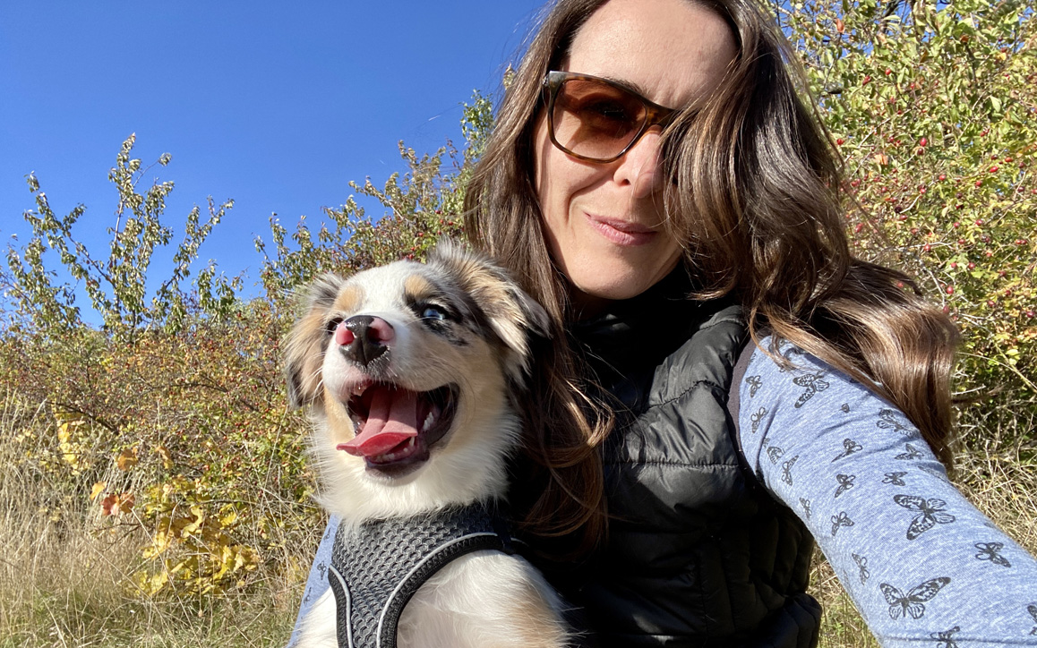 Judith Ivancsits macht ein Selfie bei einem Spaziergang in der Natur, neben ihr ein fröhlicher Hund mit Brustgeschirr.