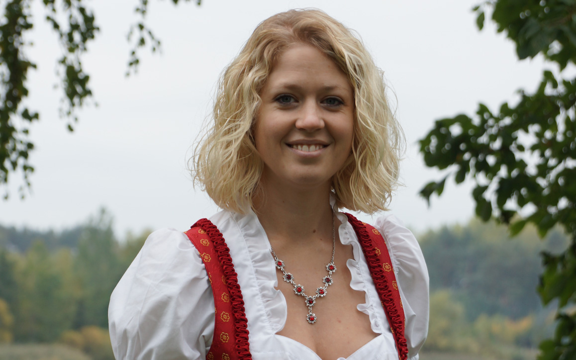 Julia Silberbauer im traditionellen Dirndlkleid, lächelnd im Freien mit Bäumen im Hintergrund.