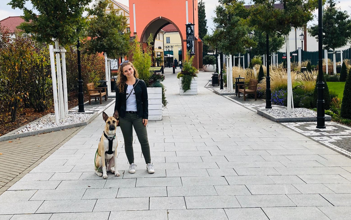 Lea Lechler steht in legerer Kleidung mit ihrem Hund „Tao“ auf einem gepflasterten Weg in einer modernen Stadtumgebung. Im Hintergrund sind Bäume, Sitzbänke und ein markantes rotes Tor zu sehen.