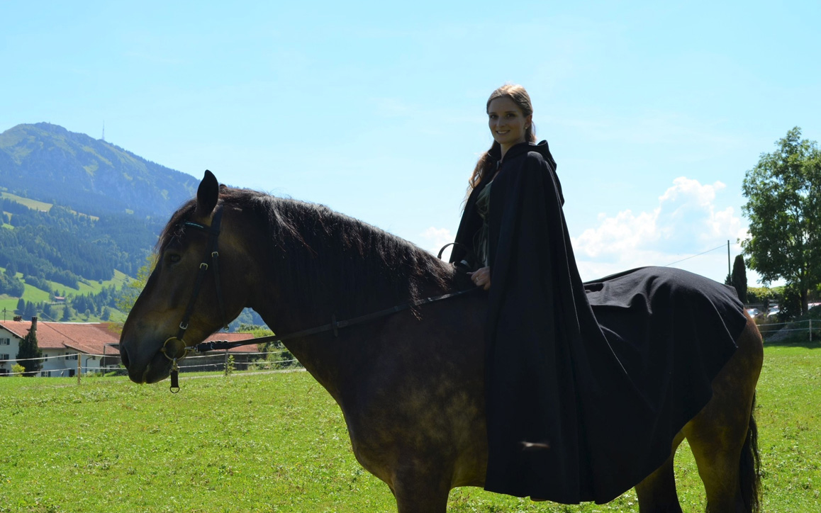 Lea Lechler sitzt in einem langen, schwarzen Mantel auf einem braunen Pferd auf einer grünen Wiese mit Bergen im Hintergrund. Sie blickt ruhig in die Kamera.