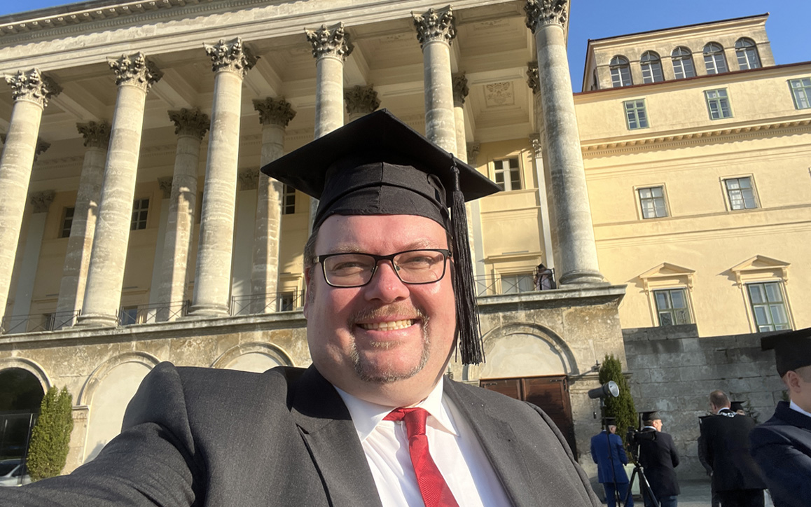 Marc Schachteli bei seiner Sponsionsfeier vor Schloss Esterházy, trägt Absolventenhut, hält ein Selfie fest
