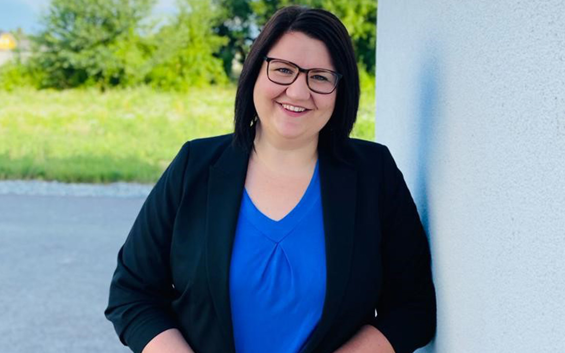 Johanna Prodinger, Studentin im MBA-Lehrgang, steht mit einem Lächeln an eine helle Wand gelehnt. Sie trägt eine Brille, ein blaues Oberteil und einen dunklen Blazer. Im Hintergrund ist eine grüne Wiese sichtbar.