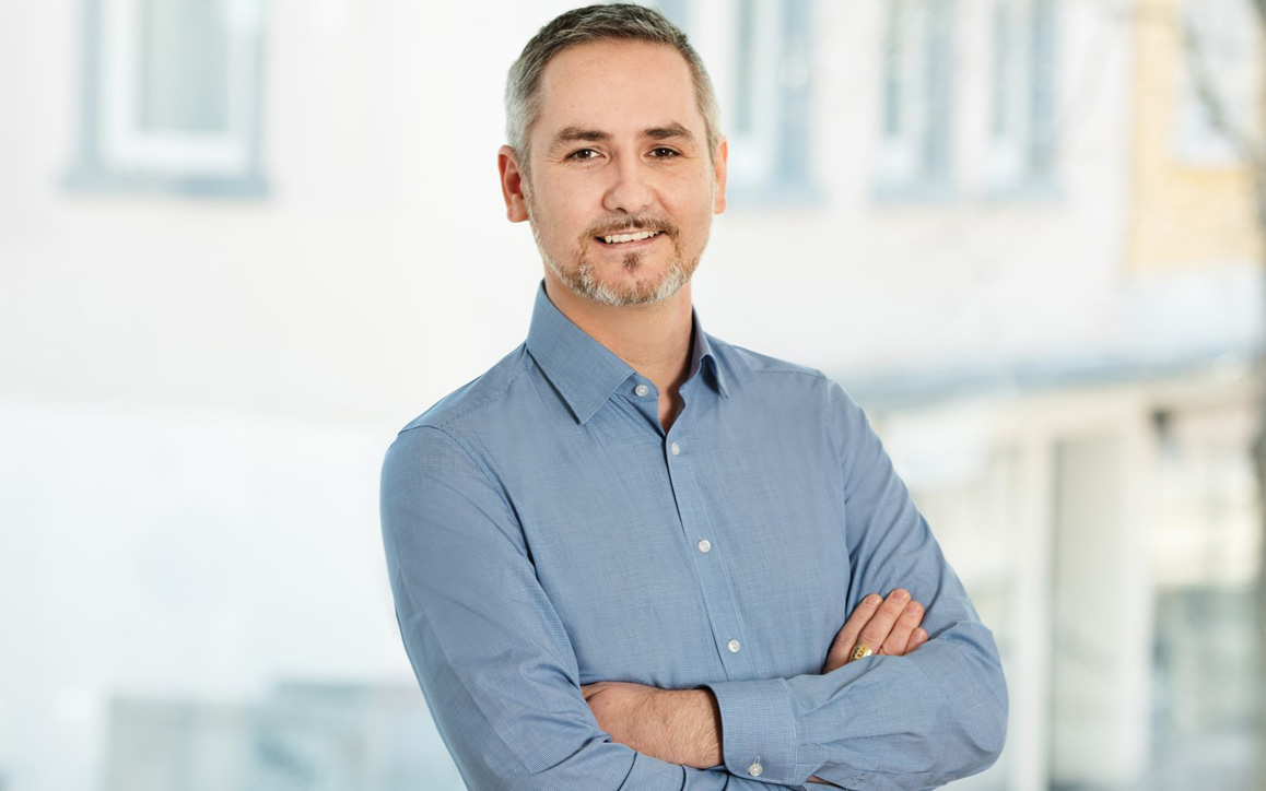 Marcel S., MBA-Student an der FH Burgenland, steht mit verschränkten Armen in einem modernen, hellen Innenraum und lächelt in die Kamera.