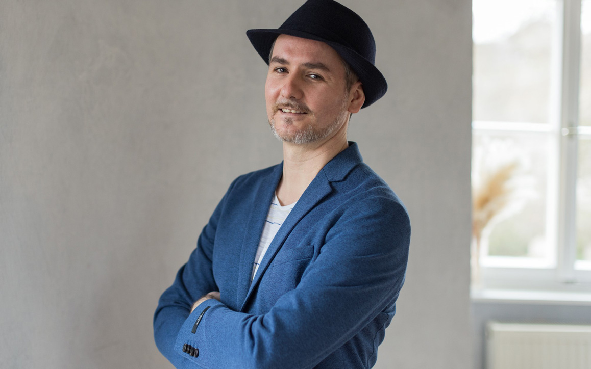 Marcel S., MBA-Student an der FH Burgenland, steht mit verschränkten Armen und dunklem Hut vor einer hellen Wand und lächelt selbstbewusst in die Kamera.