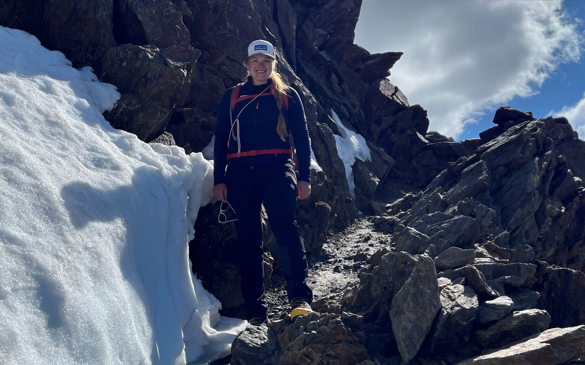 Maxi Müller beim Wandern im Hochgebirge. Ausgerüstet mit Helm, Kletterhose und Rucksack steht sie auf felsigem Untergrund zwischen Schneefeldern – Sinnbild für Ausdauer und Balance zwischen Studium und Freizeit.