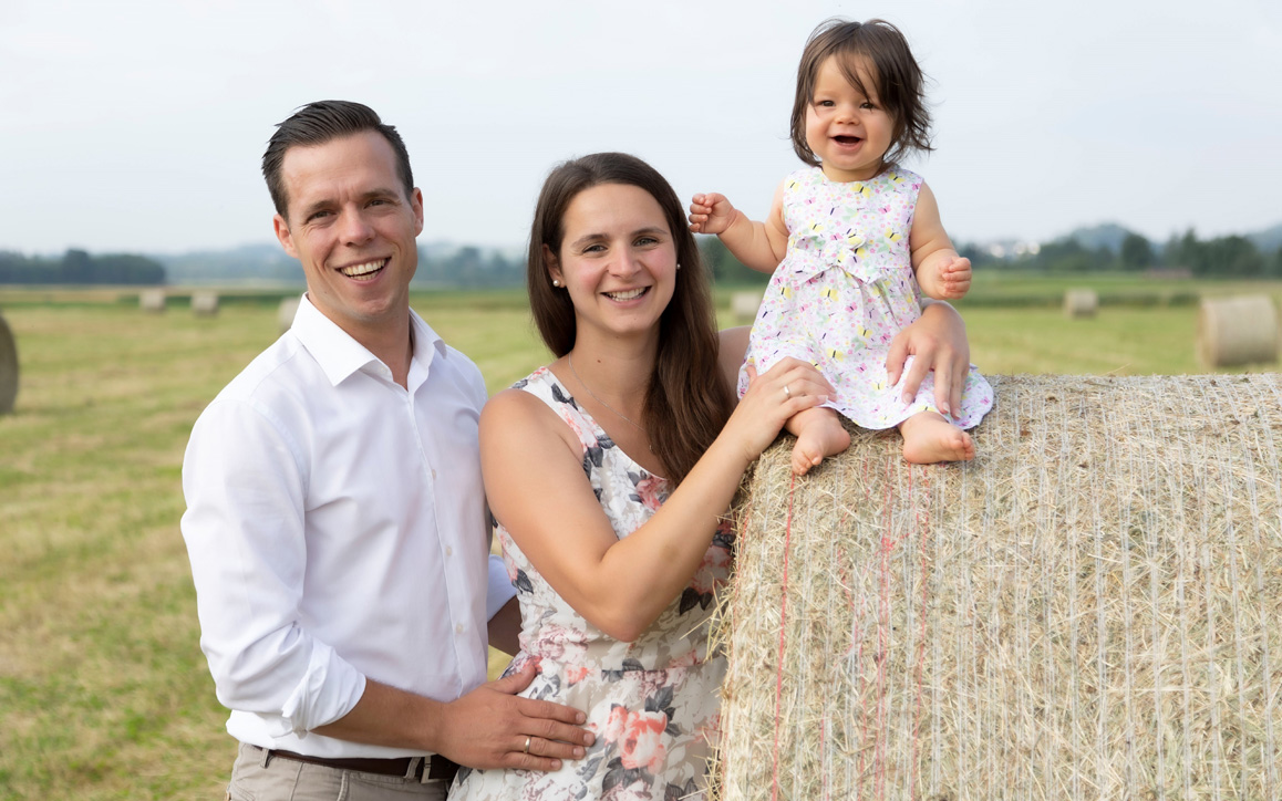 Patrick Söndgen mit seiner Partnerin und Tochter auf einem Strohballen im Grünen. Die Familie strahlt in die Kamera.
