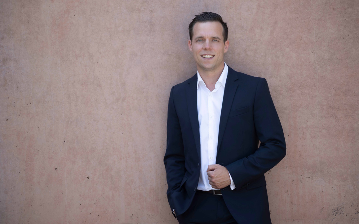Patrick Söndgen im Anzug steht entspannt vor einer rosa Betonwand, die Hände locker gefaltet, mit zuversichtlichem Blick.