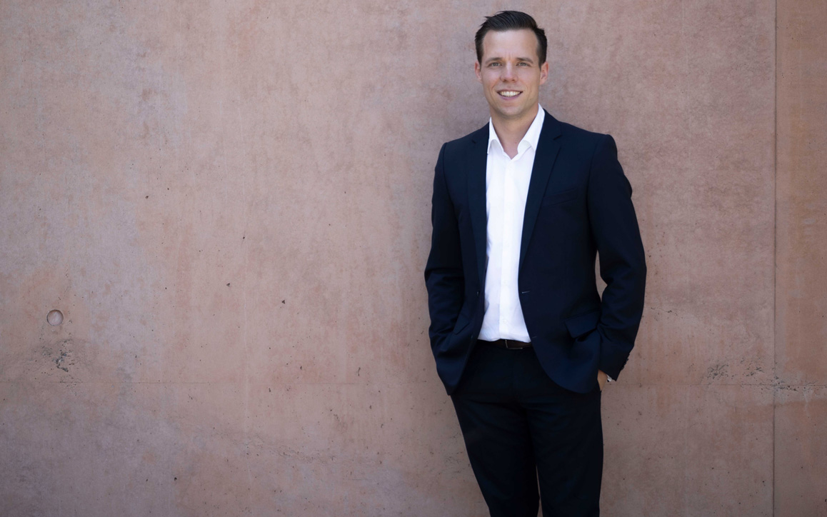 Patrick Söndgen, MBA-Alumnus der FH Burgenland, steht lässig im dunklen Anzug vor einer Betonwand und lächelt in die Kamera.