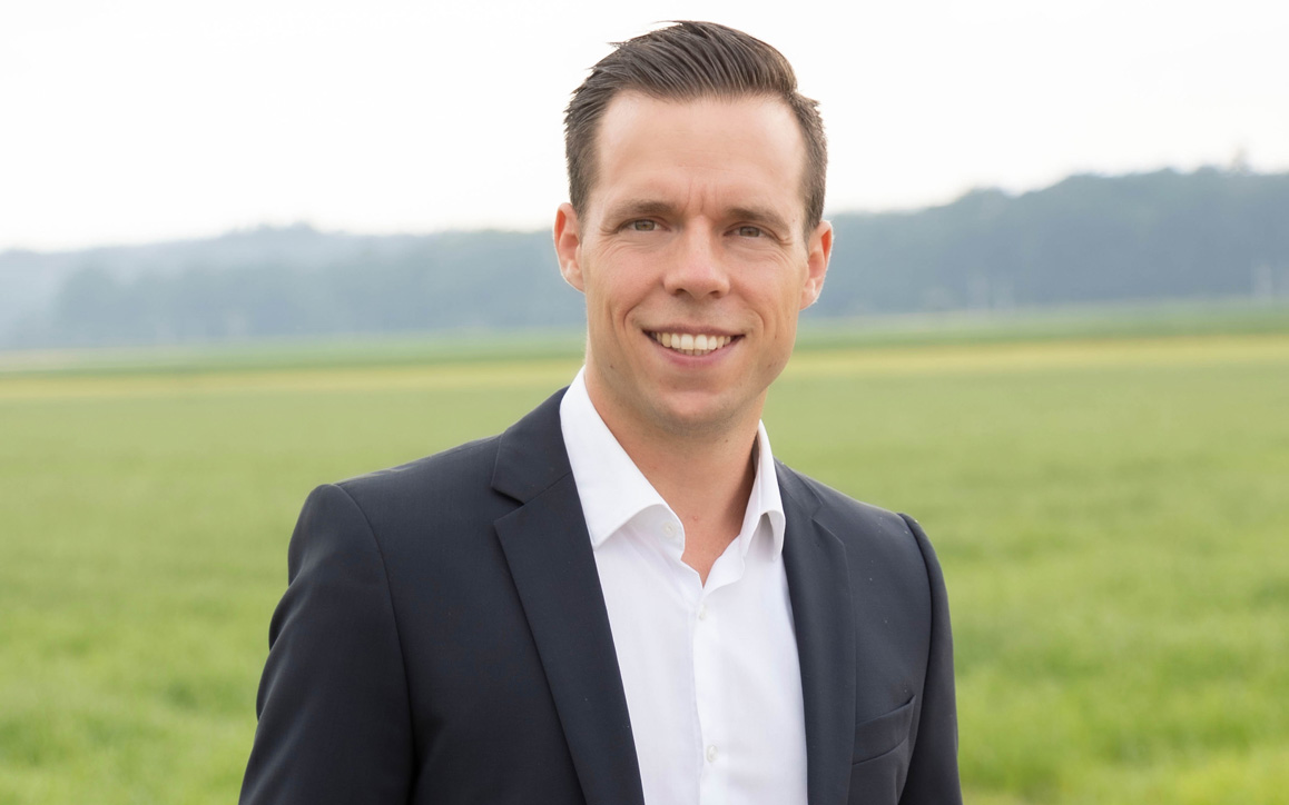 Patrick Söndgen in einem dunklen Anzug mit weißem Hemd vor einem weiten, grünen Feld. Er lächelt freundlich in die Kamera.