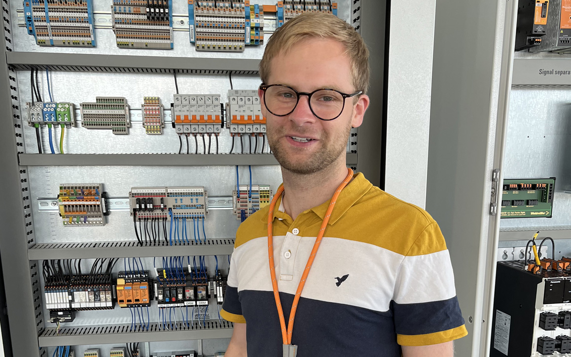 Christopher Hillebrand steht vor einer geöffneten Schalttafel mit zahlreichen technischen Komponenten und Kabeln – Einblick in sein berufliches Umfeld.