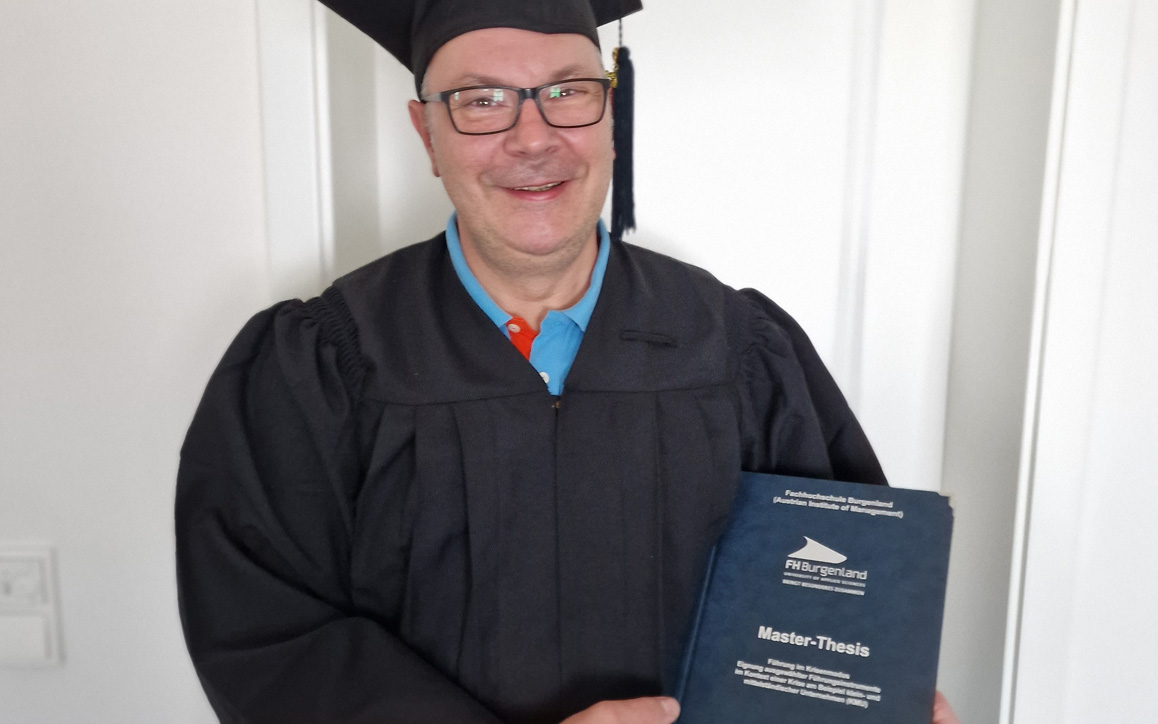Erwin Hesser trägt Talar und Abschlusskappe und hält stolz seine Master-Thesis der FH Burgenland in der Hand. 