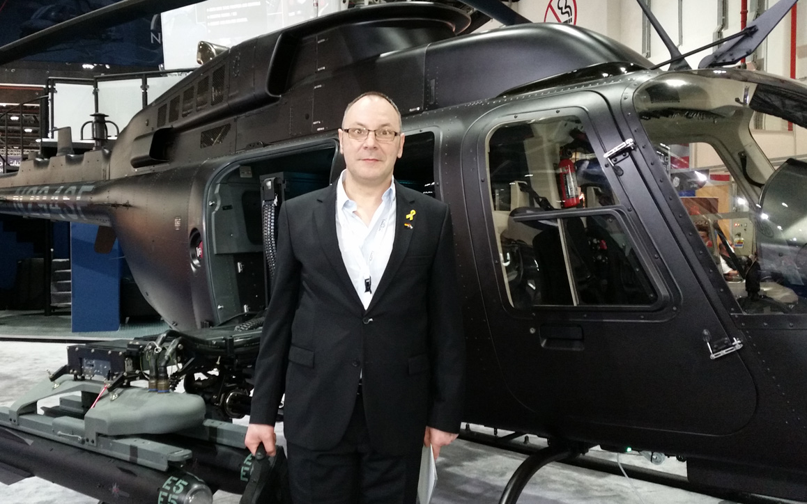 Erwin Hesser steht in einem Hangar vor einem militärischen Helikopter. Er trägt einen dunklen Anzug mit Anstecker und schaut direkt in die Kamera.