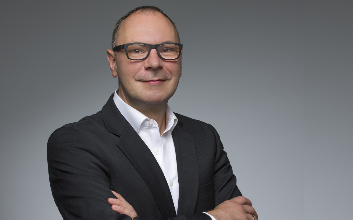 Erwin Hesser, MBA-Absolvent der FH Burgenland, blickt selbstbewusst in die Kamera. Er trägt einen dunklen Anzug und eine Brille, der Hintergrund ist neutral grau gehalten.