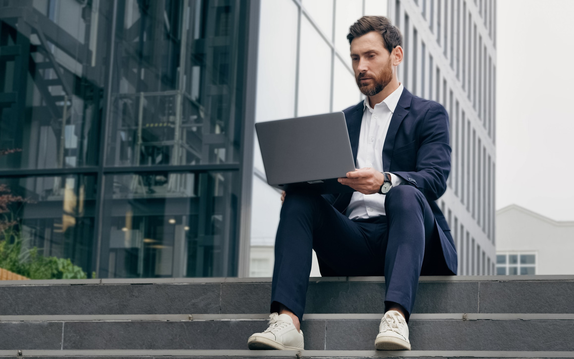 Junger Mann im Business-Outfit sitzt mit Laptop auf einer Außentreppe vor einem modernen Bürogebäude – symbolisch für flexibles Fernstudium an der Hochschule Burgenland.