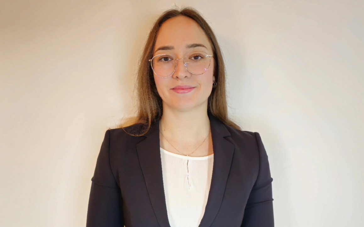 Anna Panzenböck steht vor einer neutralen Wand und trägt einen dunklen Blazer über einer weißen Bluse. Ihre Brille und ihr dezentes Lächeln verleihen dem Bild einen selbstbewussten, professionellen Ausdruck.