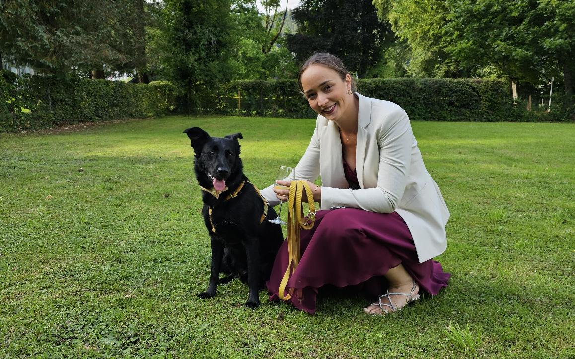 Anna Panzenböck kniet auf einer gepflegten Grünfläche und hält freundlich lächelnd ihren schwarzen Hund an der Leine. Sie trägt ein elegantes, bordeauxfarbenes Kleid und einen weißen Blazer.