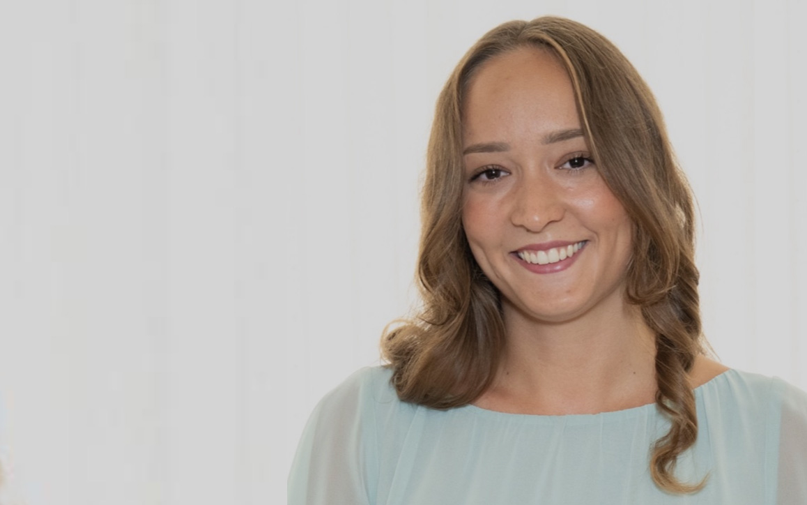 Anna Panzenböck lächelt freundlich in die Kamera. Sie trägt ein helles Oberteil und hat schulterlanges, gewelltes Haar. Der neutrale Hintergrund unterstreicht ihren professionellen Auftritt.