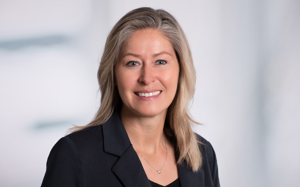 Porträt von Anja Löschmann vor neutralem grauen Hintergrund, freundlich lächelnd, in dunklem Blazer.