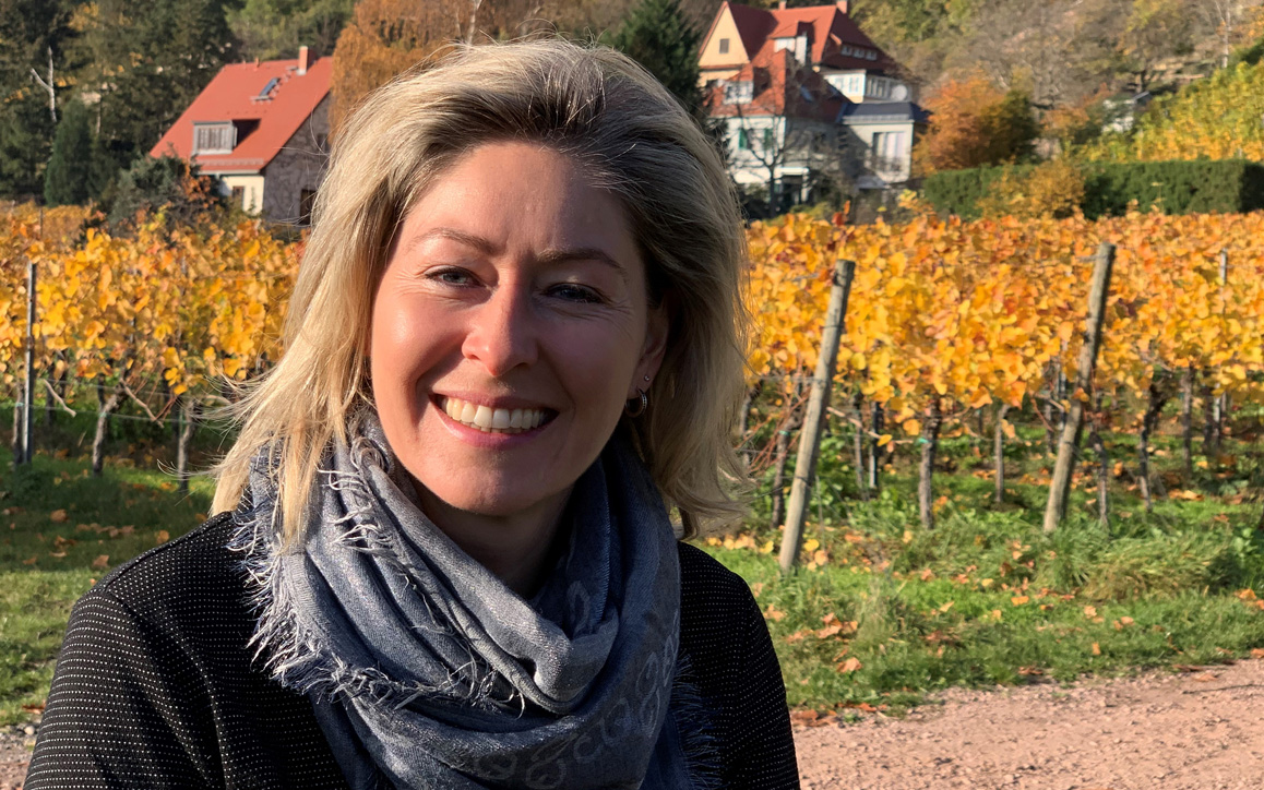 Anja Löschmann in herbstlicher Landschaft mit Weingarten im Hintergrund, sie lächelt in die Kamera und trägt einen grauen Schal.