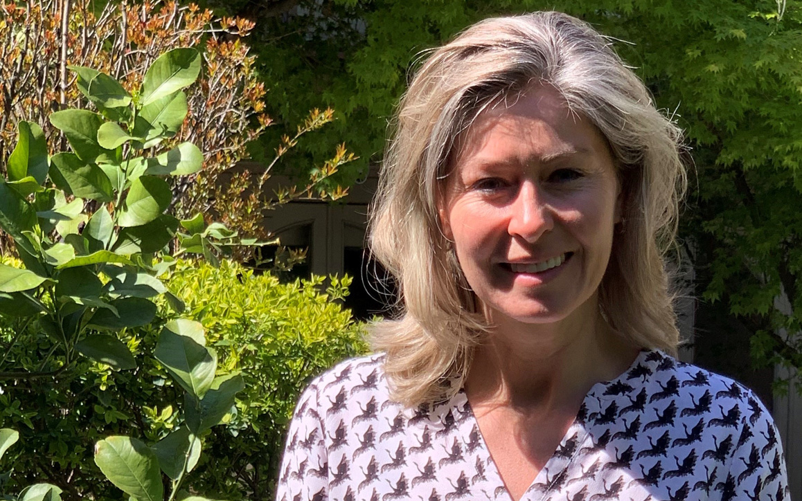 Anja Löschmann steht in einem Garten, umgeben von grüner Vegetation, und lächelt, bekleidet mit einer hellen Bluse mit Vogelmuster.