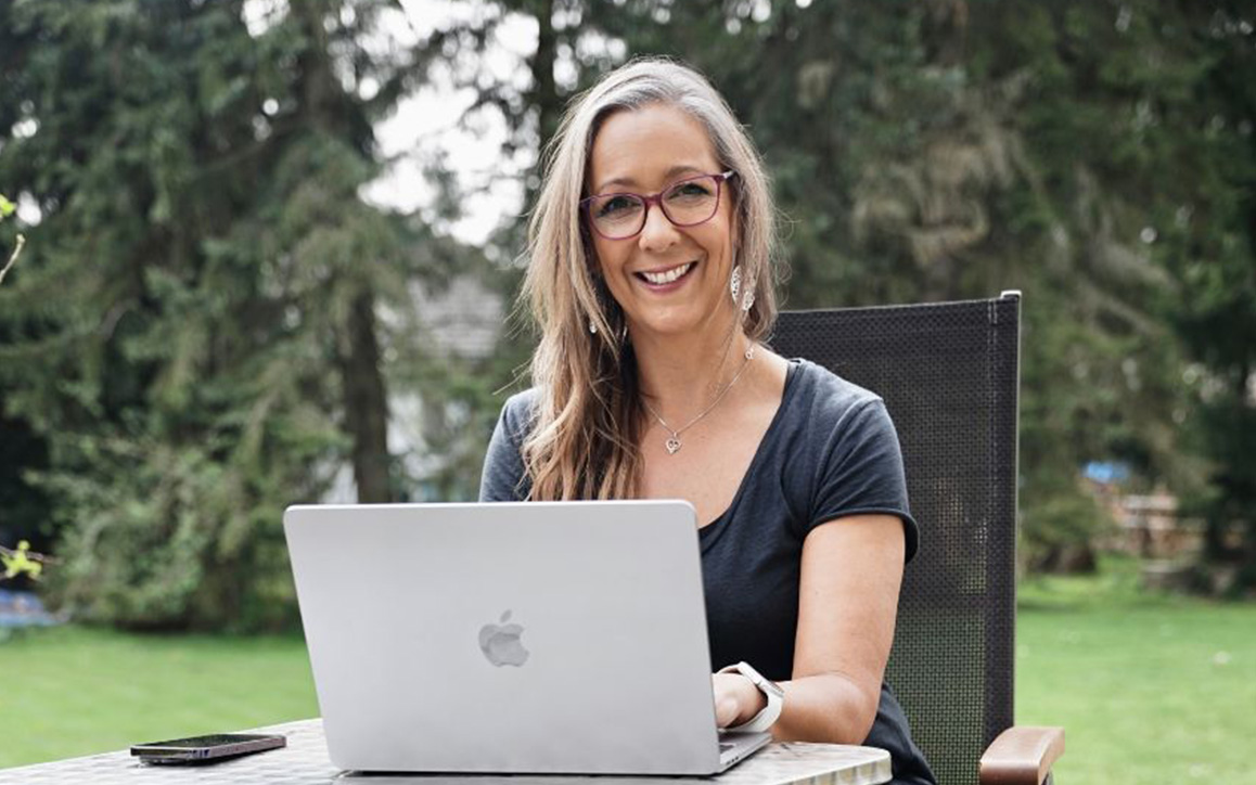 Elke Egi sitzt entspannt im Garten an einem Tisch und arbeitet an einem silbernen Laptop – ein Symbol für Flexibilität und Selbstbestimmung im Fernstudium.