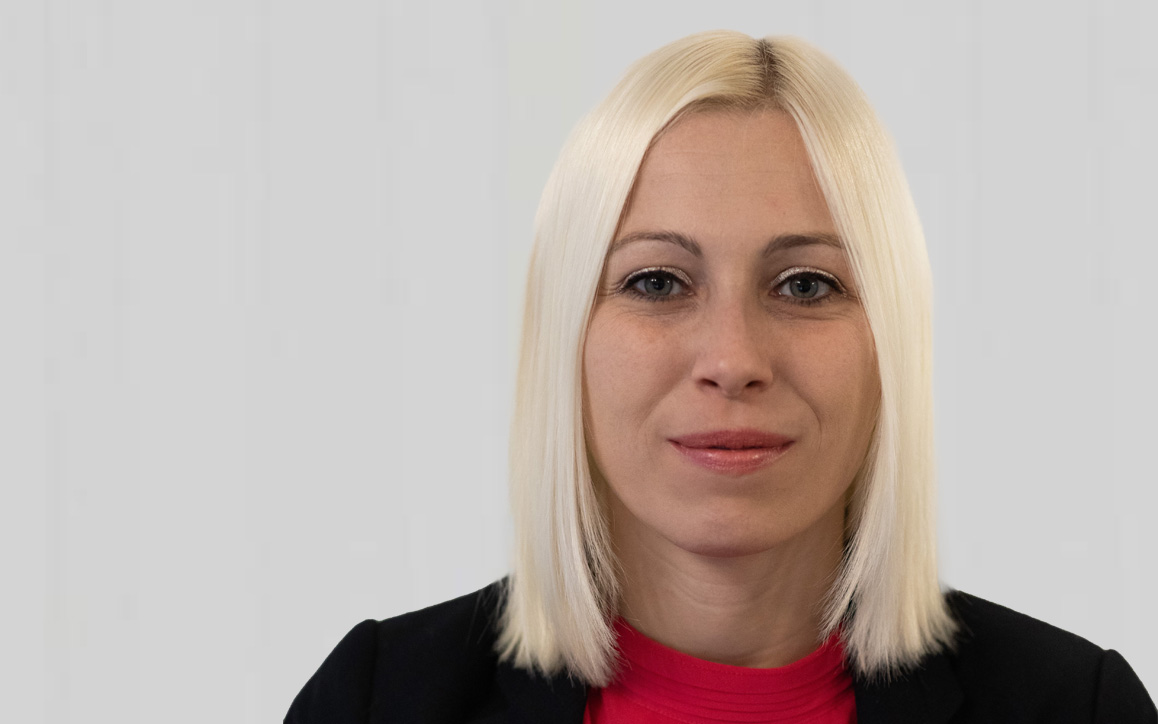 Porträt von Manuela Schoyswohl, MBA-Absolventin der FH Burgenland, mit blondem Haar, schwarzem Blazer und rotem Shirt, vor hellem Hintergrund.