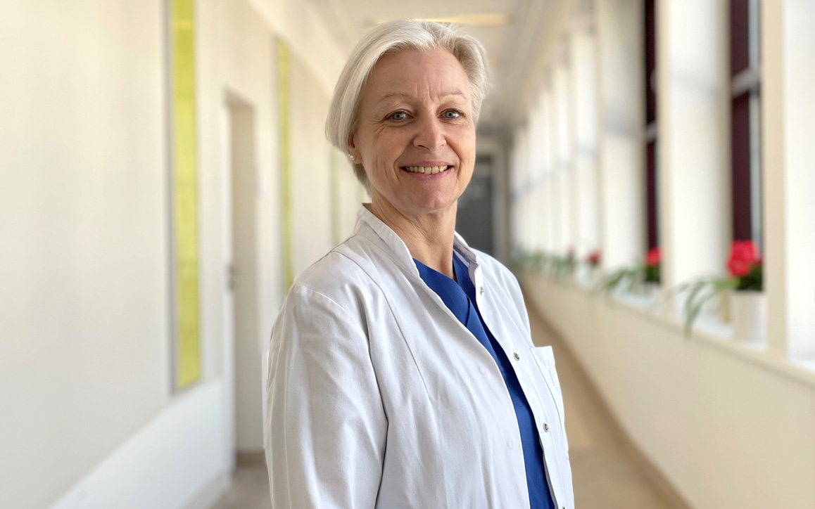 Birgit Giese im weißen Kittel lächelt in einem hellen Klinikflur in die Kamera.