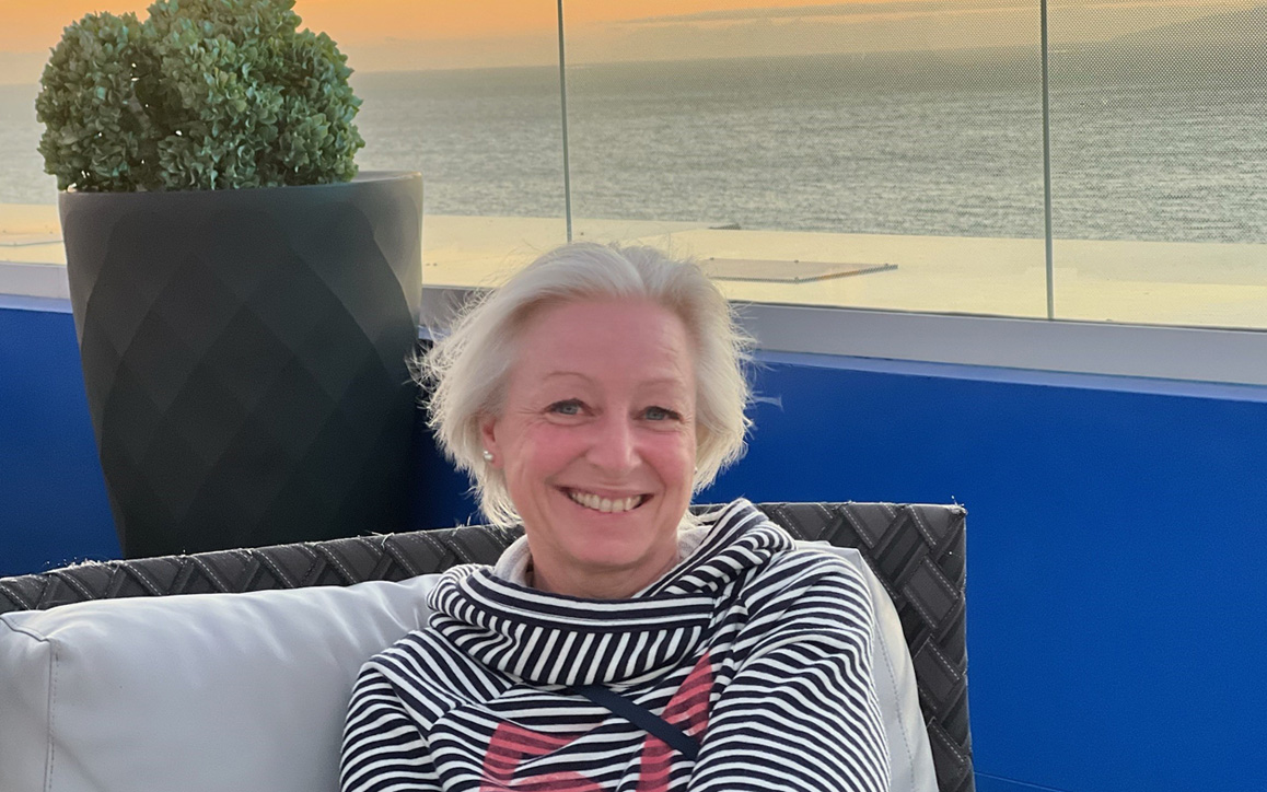 Birgit Giese sitzt entspannt in gestreiftem Pullover auf einer Terrasse mit Blick aufs Meer.