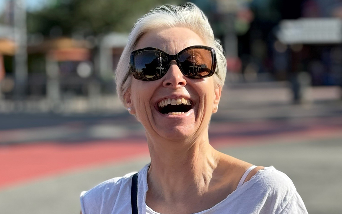 Birgit Giese mit Sonnenbrille lacht herzlich bei Sonnenschein im Freien.