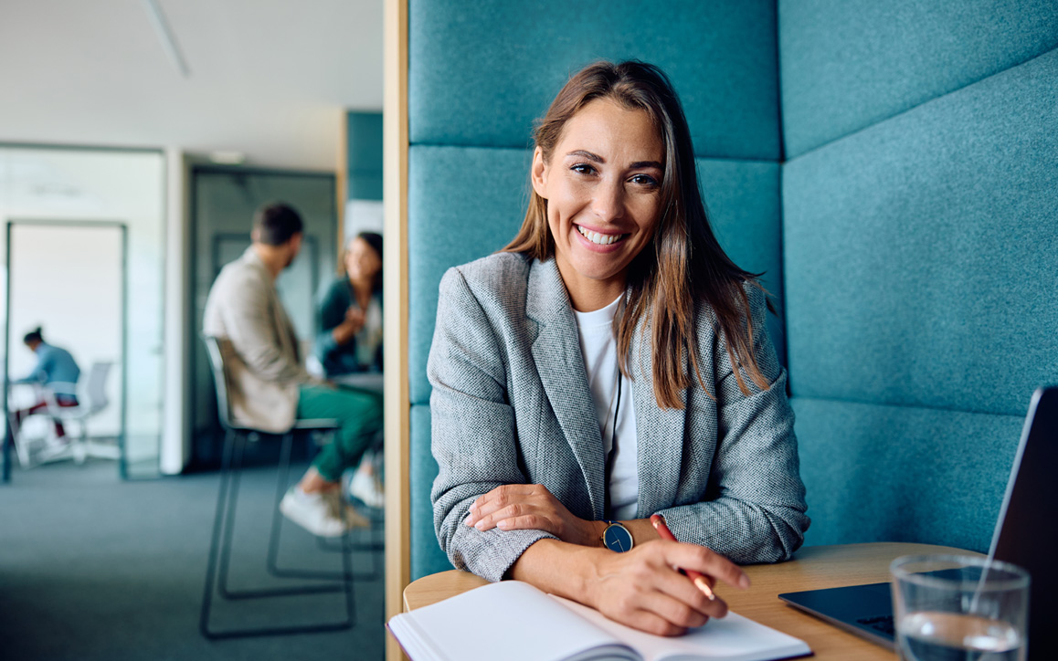 Junge Frau mit Notizbuch und Laptop sitzt lächelnd an einem Tisch in modernem Büro – steht für zeitgemäßes Arbeiten im Personalmarketing.