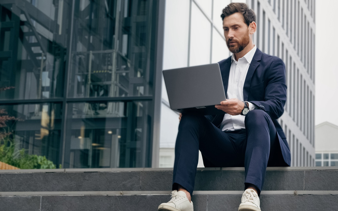 Berufsbegleitend zum Bachelor an der Hochschule Burgenland Junger Mann im Anzug sitzt mit Laptop vor einem modernen Gebäude. Das Bild steht für den berufsbegleitenden Einstieg ins Bachelor-Studium an der Hochschule Burgenland.