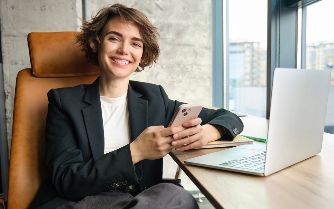 Junge Frau sitzt lächelnd am Schreibtisch mit Laptop, im Hintergrund ein helles Büro – symbolisiert persönliche Weiterentwicklung im berufsbegleitenden Fernstudium.