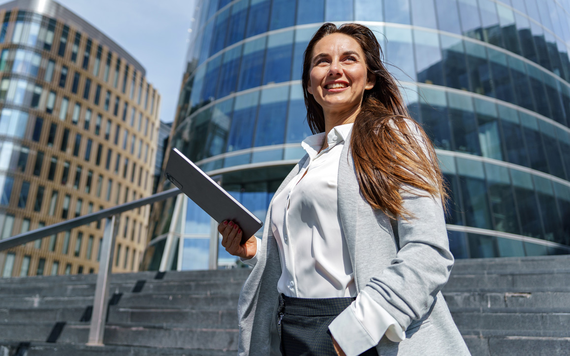 Junge Frau in Business-Outfit steht mit Tablet vor modernem Businessgebäude – symbolisiert internationale Perspektiven, unternehmerisches Denken und Diversität im globalen Management.