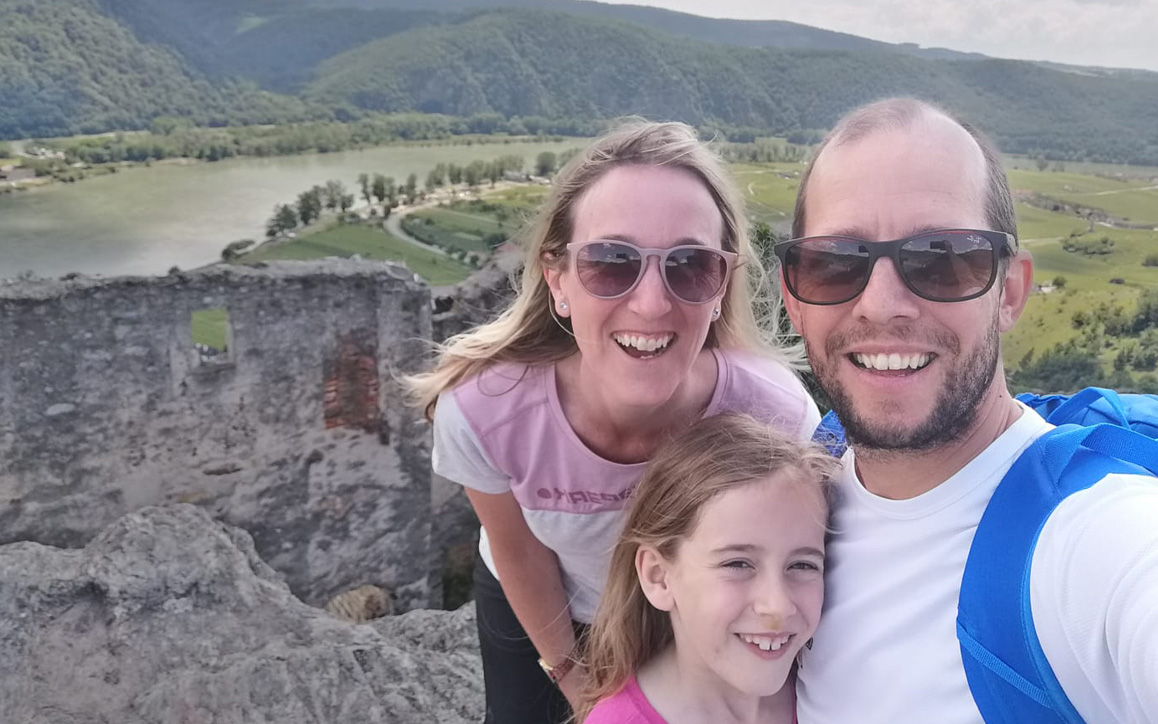 Thomas Bravo lächelt gemeinsam mit seiner Frau und Tochter in die Kamera, im Hintergrund eine malerische Landschaft mit Hügeln, Wald und Ruine.