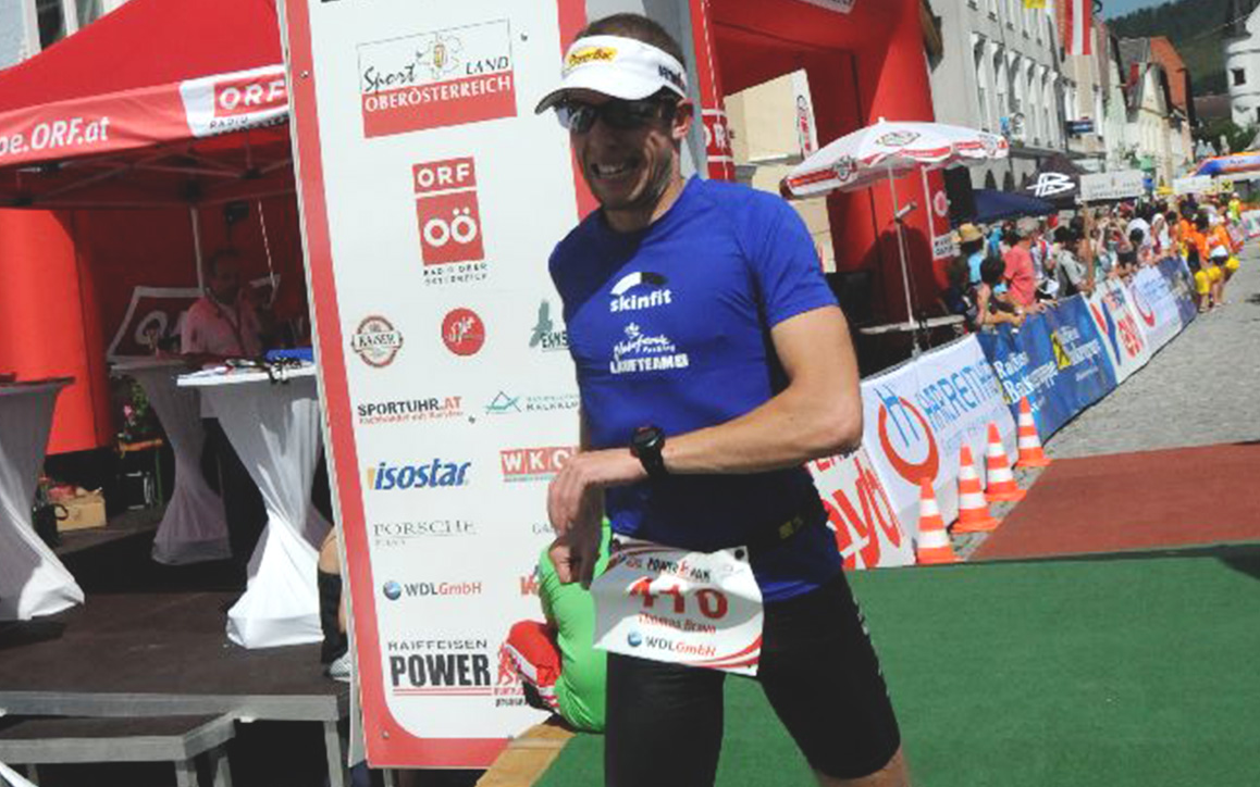 Thomas Bravo läuft mit Startnummer und Sportbekleidung durch das Ziel eines Marathons in Oberösterreich, im Hintergrund Zuschauer und Sponsorenbanner.