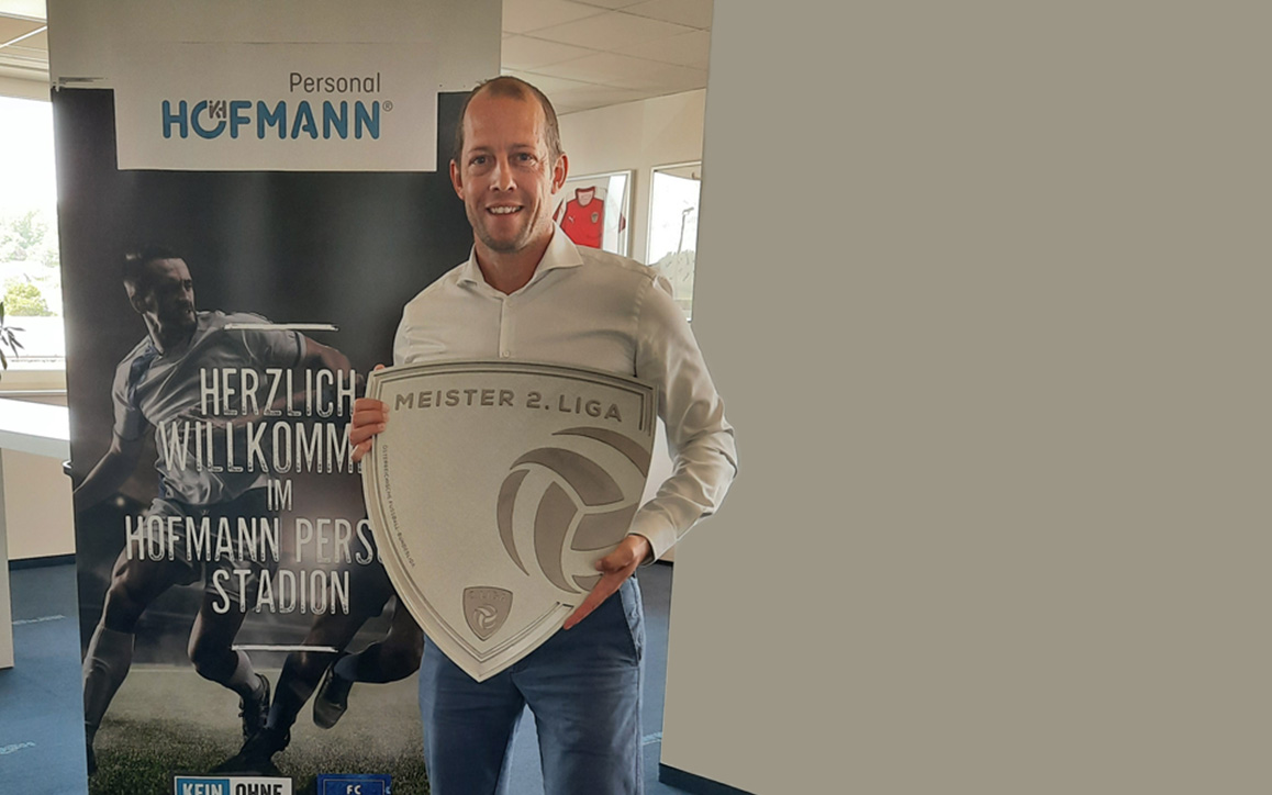 Thomas Bravo steht vor einem Roll-up mit der Aufschrift „Herzlich willkommen im Hofmann Personal Stadion“ und hält ein Schild mit dem Titel „Meister 2. Liga“ in den Händen.