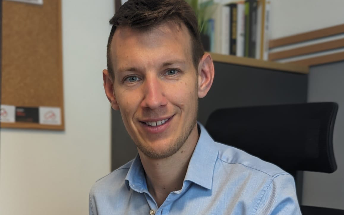 Christoph Tatzber, MBA-Absolvent der Hochschule Burgenland, sitzt lächelnd an einem Schreibtisch in einem Büro.